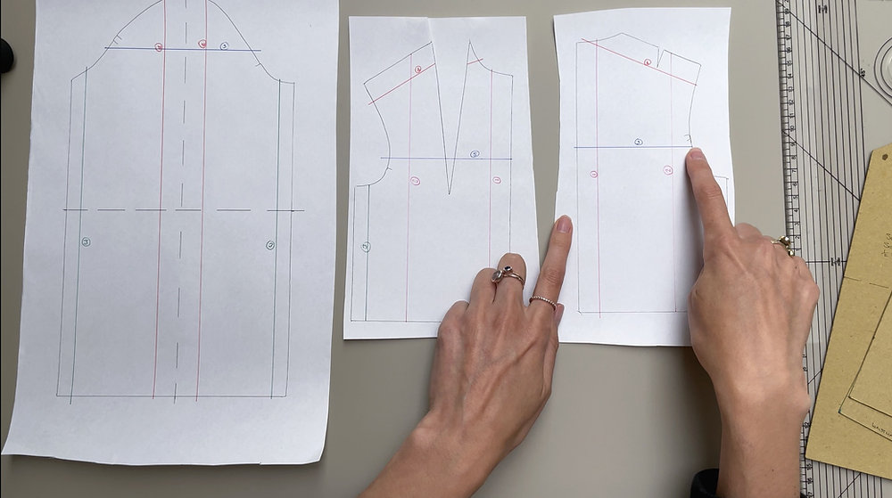 This image shows three separate garment pattern pieces laid out on a flat surface. The pattern pieces are made from white paper with technical drawings in various colors, including blue, red, and black lines, indicating garment construction lines and measurements. Two hands with rings are pointing at specific sections on the middle and right pattern pieces. The left pattern piece is larger and more rectangular, likely representing a back or front bodice piece, while the middle and right pieces are smaller with distinct shapes typical of garment sections such as the front bodice and yoke or collar. In the background, parts of a ruler and brown pattern paper with additional markings can be seen. The image is useful for illustrating garment pattern drafting, sewing tutorials, or fashion design documentation.