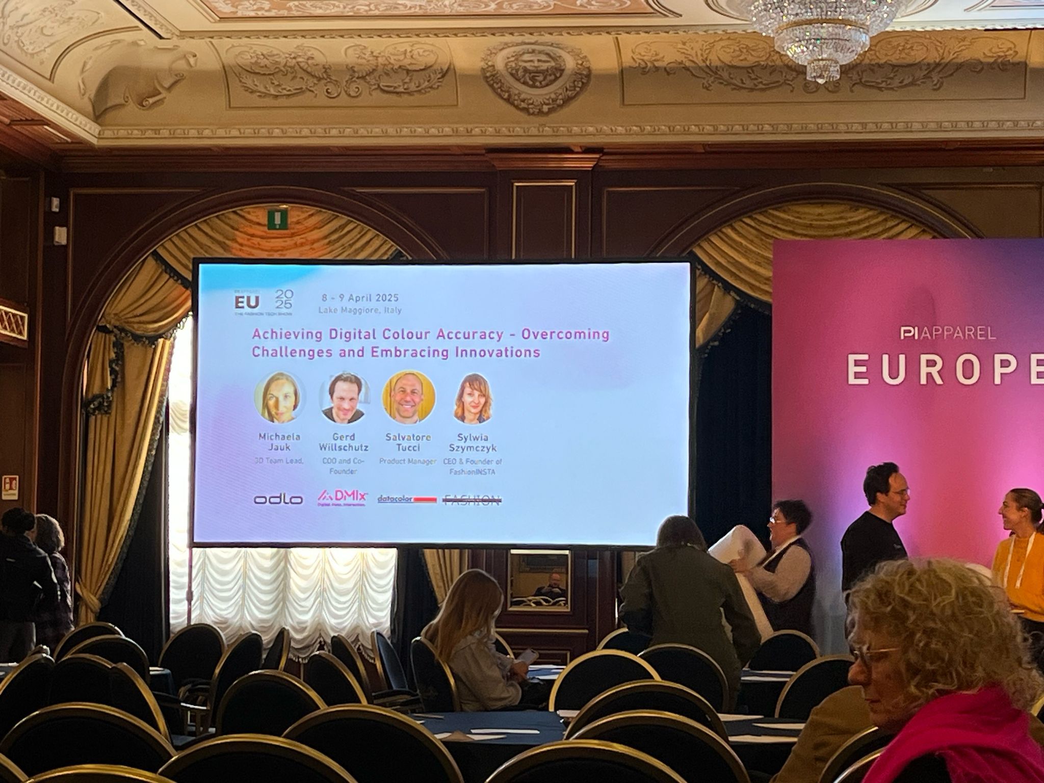 The image shows a conference room setup for a panel discussion titled 'Achieving Digital Colour Accuracy - Overcoming Challenges and Embracing Innovations,' scheduled for 8-9 April 2025 in Lake Maggiore, Italy. A large screen displays the event title along with photos and names of the four panelists: Michaela Jauk (3D Team Lead), Gerd Willschutz (COO and Co-Founder), Salvatore Tucci (Product Manager), and Sylwia Szymczyk (CEO and Founder of FashionINSTA). The session appears to be part of the PI Apparel Europe 2025 event. The room features rows of chairs and attendees preparing for the session, highlighting a professional setting focused on digital fashion technology and innovation.
