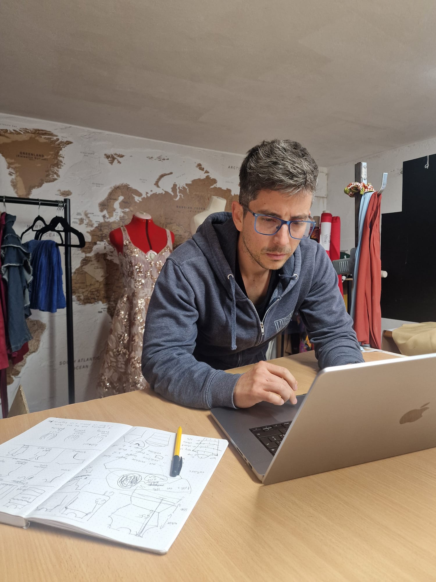 The image shows a male fashion designer focused on his laptop in a well-lit workspace. In front of him on the wooden table lies an open notebook filled with detailed technical sketches and notes related to garment pattern making alongside a yellow pen. Behind him, a rack with various fabrics and a mannequin dressed in a detailed embellished dress can be seen, indicating a creative fashion design studio environment. The background wall features a world map, enhancing the professional and global design atmosphere. This image depicts the process of digital and manual pattern making in fashion design.
