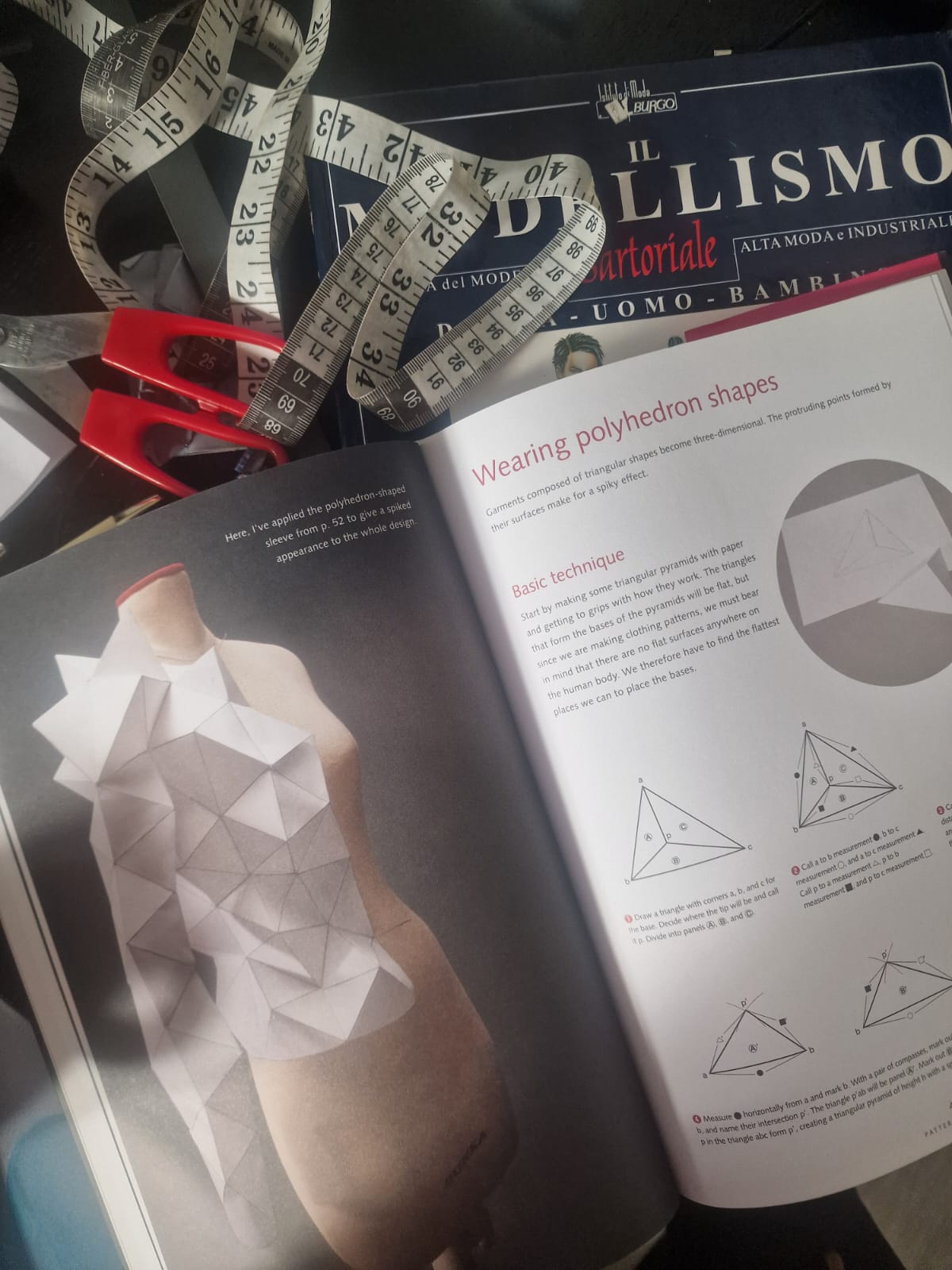 This image shows an open fashion design book page explaining the technique of creating garments composed of triangular polyhedron shapes to achieve a spiky 3D effect. On the left, a dress form is shown with a garment featuring a polyhedron-shaped sleeve made from paper, demonstrating the design concept discussed. The right page provides detailed instructions and diagrams illustrating how to draft and measure triangular pyramids for garment construction. Surrounding the book are tailoring tools such as red-handled scissors and a white measuring tape, indicating a workspace for fashion design and garment pattern making. The book in the background titled 'Il Modellismo Sartoriale' suggests the content is related to industrial and high fashion tailoring for men and children. This image is useful for professionals and students interested in advanced garment pattern creation involving geometric shapes.