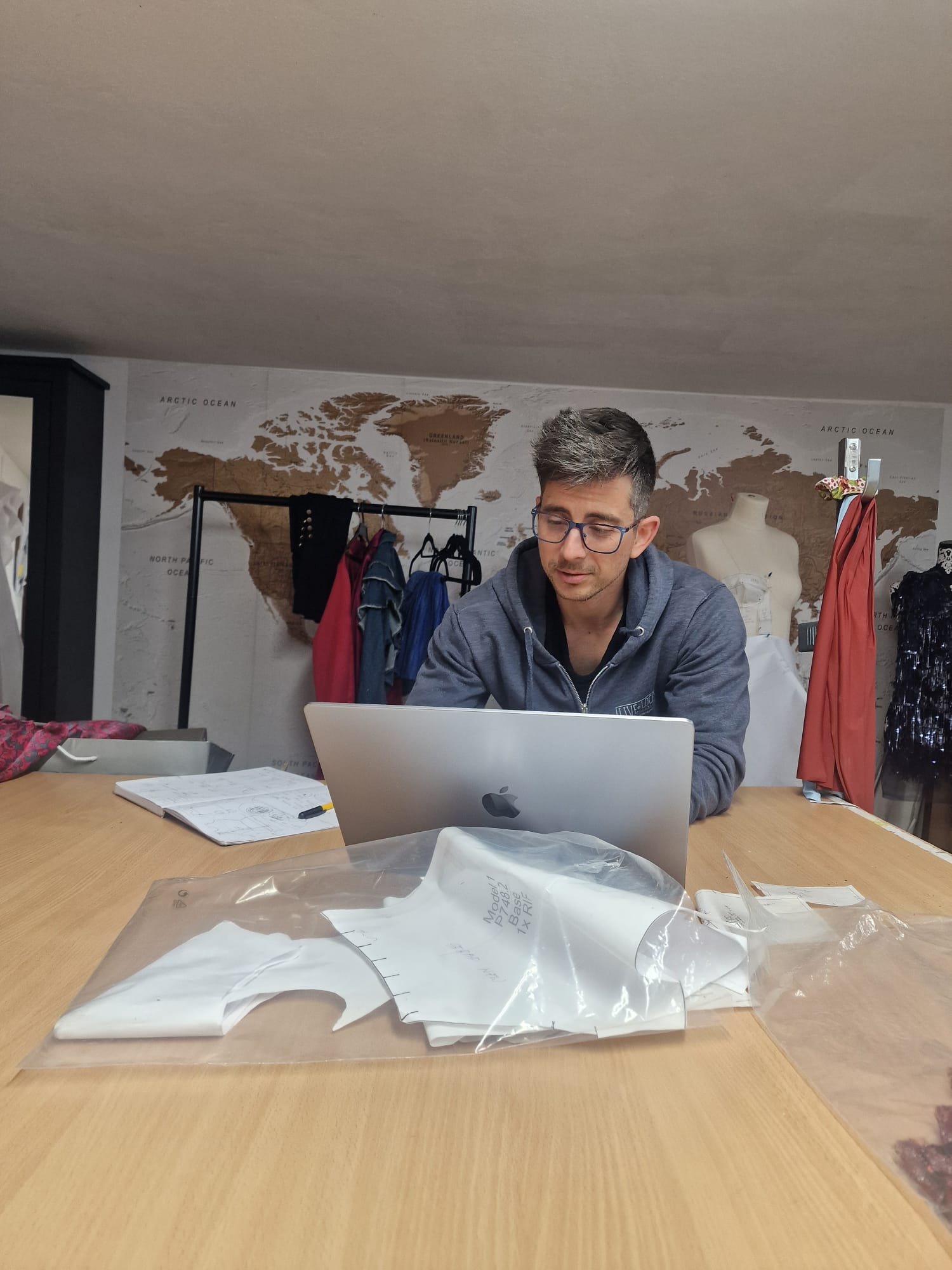 The image depicts a man working on a silver MacBook laptop at a wooden table in a fashion design studio. On the table in front of him are plastic-wrapped sewing patterns for clothing pieces. Behind him is a clothing rack with various garments and a world map mural on the wall. The studio also contains dress forms with partially completed dresses, indicating active fashion design and garment construction processes. The setting suggests a workspace environment for fashion design, pattern making, and garment development.