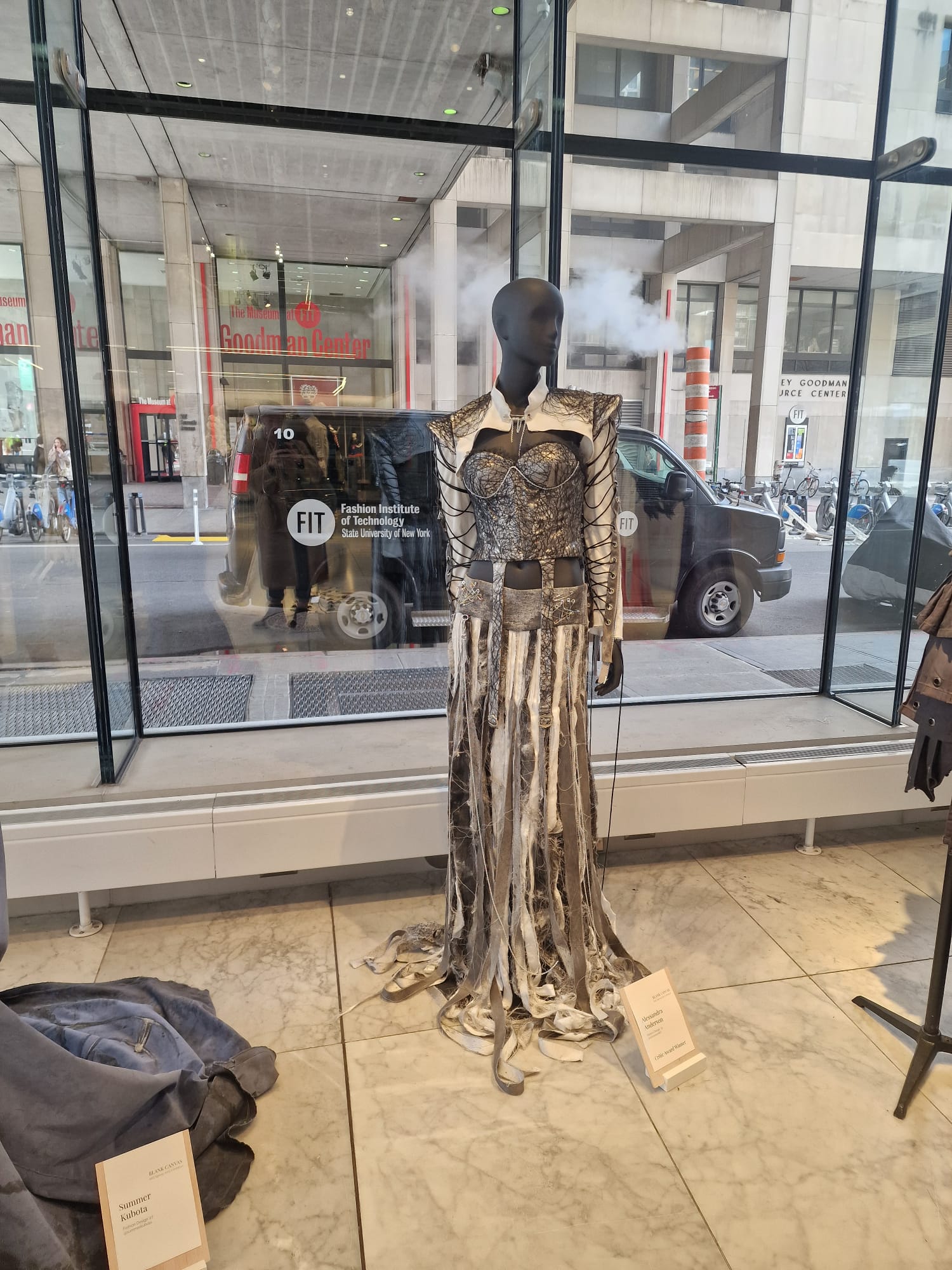 This image shows a mannequin dressed in an avant-garde fashion outfit displayed inside a glass showroom at the Fashion Institute of Technology (FIT), State University of New York. The fashion piece includes a structured top with intricate web-like detailing and long, flowing strips of fabric extending from the waist down, creating a dramatic skirt effect. The showroom has large glass windows through which the street and a FIT vehicle are visible. Placards with designer names and collection titles are situated near the garments, indicating a curated exhibit of experimental or student fashion design projects. The setting suggests a professional environment for showcasing innovative fashion concepts.
