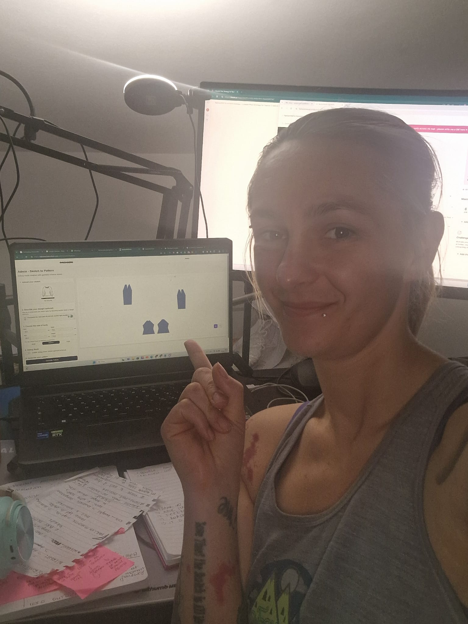 The image shows a woman smiling and pointing towards the screen of a laptop on her desk. The laptop screen displays a software interface labeled "Admin - Sketch to Pattern," which features digital fashion sketches of garment pieces. The woman appears to be in a casual workspace, with a microphone boom arm visible in the background, as well as notes and papers scattered on the desk. The setting suggests a workspace used for fashion design or development, demonstrating the use of digital tools for converting sketches into sewing patterns.