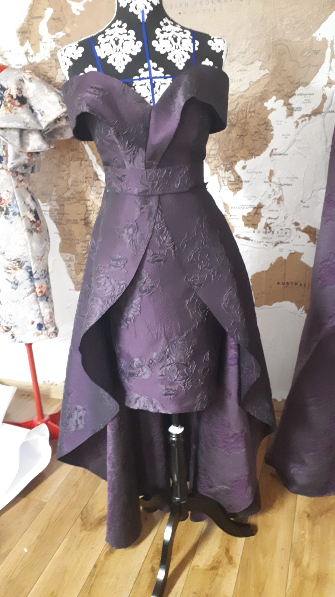 The image shows a stylish purple off-shoulder dress displayed on a black and white patterned mannequin. The dress features a high-low hemline, elaborate floral jacquard fabric texture, and a fitted bodice with a structured neckline. The setting appears to be a design studio or showroom, with a large world map on the wall in the background and additional fabric and dress forms nearby. This dress could be used for evening wear or special occasions and is an example of contemporary cocktail or formal dress design.