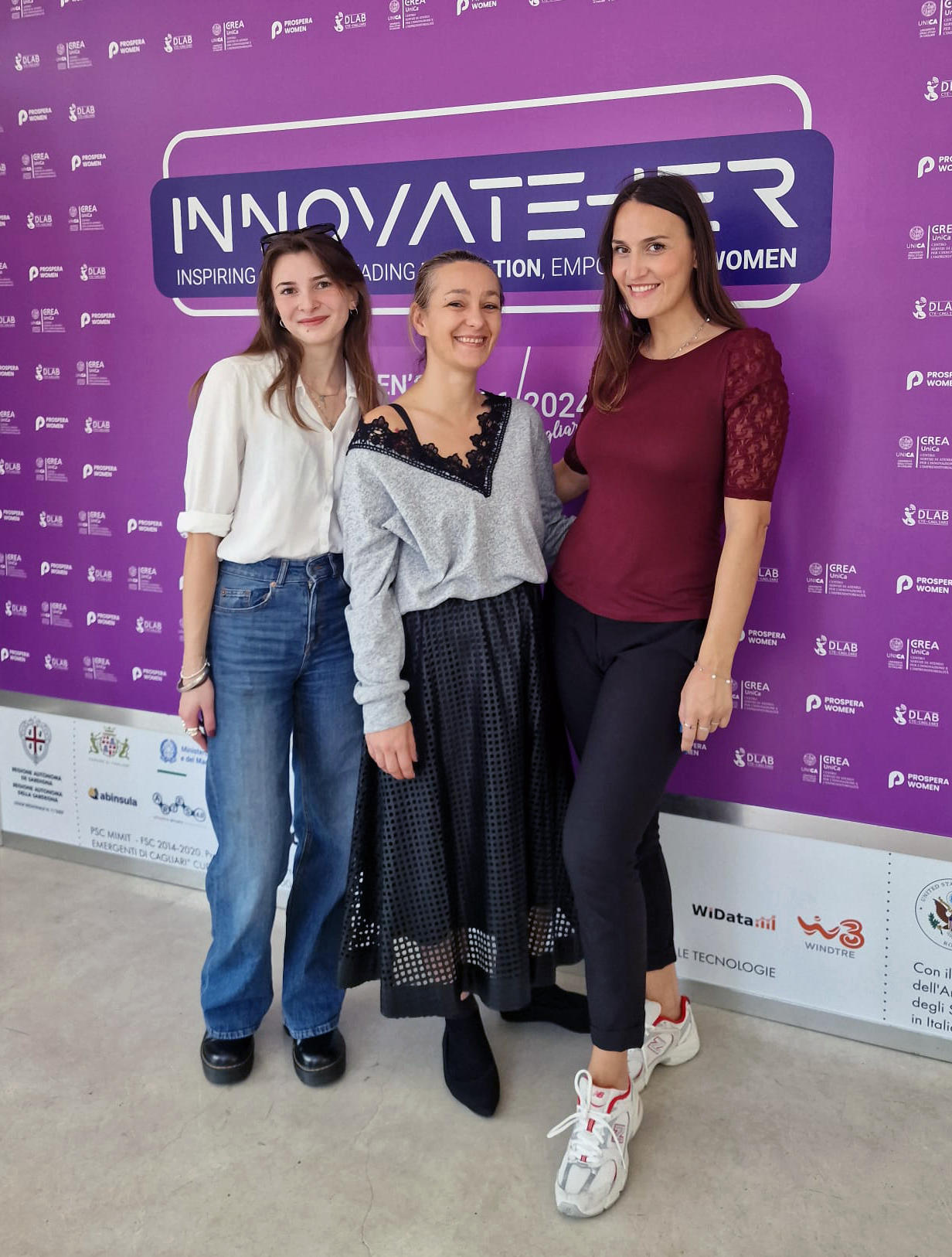 This image shows three women standing and smiling in front of a purple backdrop for the InnovateHER event in 2024. The backdrop features the InnovateHER logo and the tagline 'Inspiring, Leading, Inclusion, Empowerment of Women.' The background also includes logos of various sponsors and partners such as Prospera Women, DLAB, and others. The setting appears professional and empowering, highlighting the theme of women's leadership and innovation. The image is suitable for use in documentation or knowledge-sharing contexts related to women in business, innovation, empowerment events, and networking.