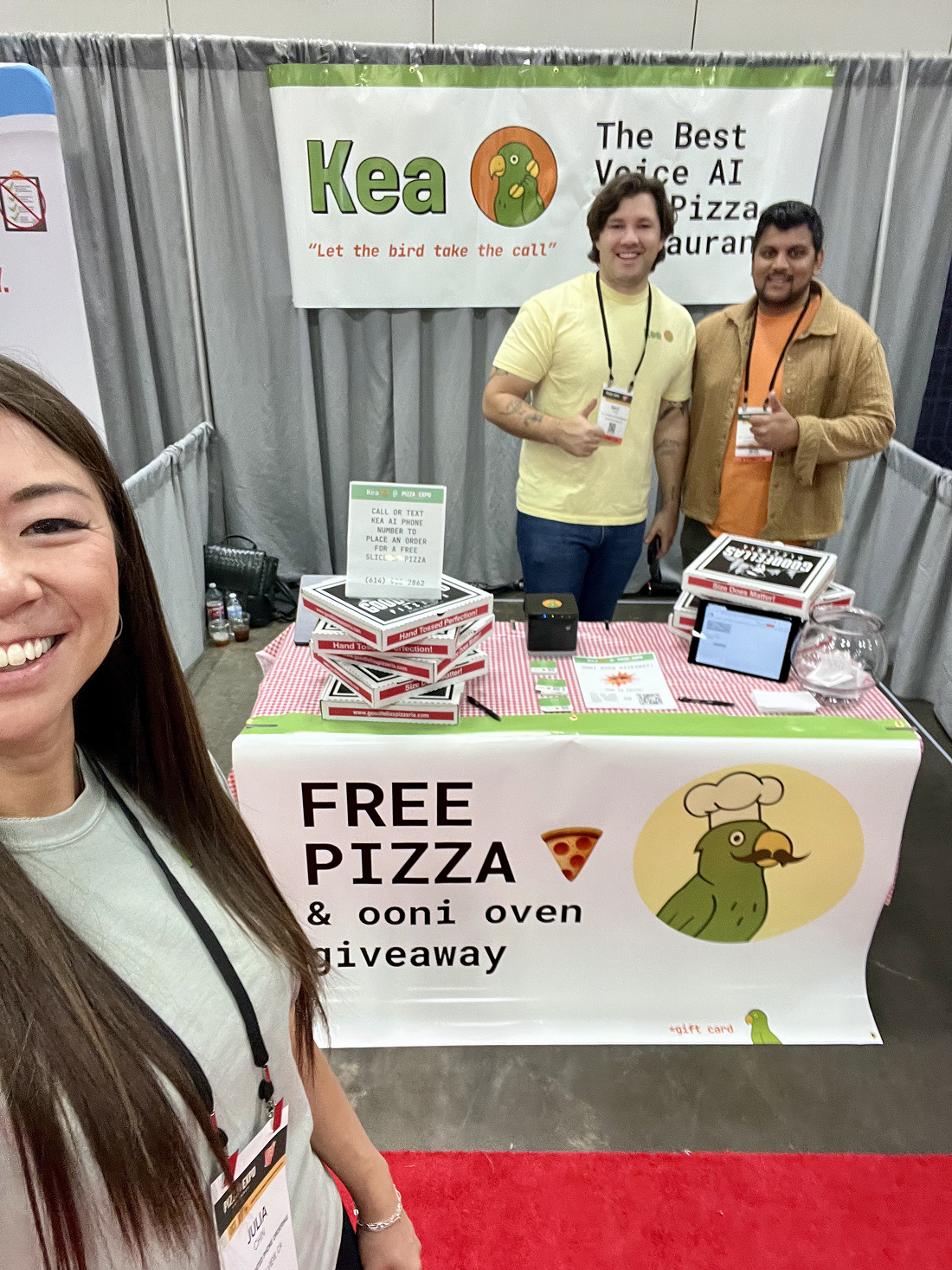 This image shows three members of Team Kea at their booth during the Pizza Expo event. The booth features signage promoting Kea as "The Best Voice AI for Pizza Restaurants" with the slogan "Let the bird take the call." The table is decorated with pizza boxes and promotional material offering free pizza slices and an Ooni oven giveaway. Two team members stand behind the table giving thumbs-up, while a third person takes a selfie. This image illustrates Kea's marketing efforts and engagement strategy at a trade show focused on pizza industry professionals.