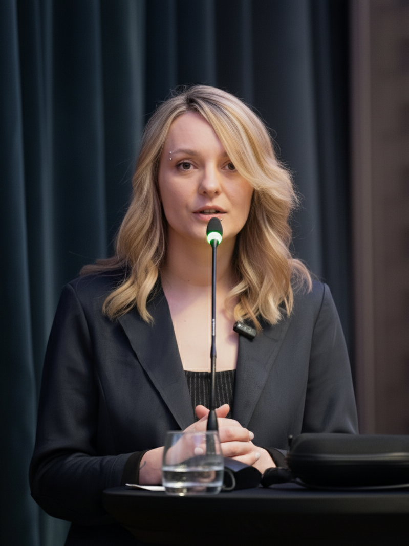 The image depicts a professional woman with blonde hair styled in loose waves, speaking at a conference podium. She is wearing a dark blazer over a black top and addressing an audience through a microphone on a stand. In front of her on the podium is a glass of water and some documents. The background features a dark curtain, suggesting an indoor professional setting. This image is suitable for use in business presentations, corporate profiles, or articles on leadership and public speaking.